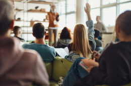 Back view of female student raising her hand to ask a question while having a class in a casual classroom.