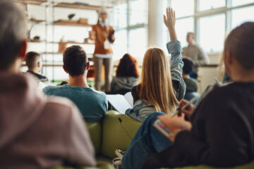 Back view of female student raising her hand to ask a question while having a class in a casual classroom.